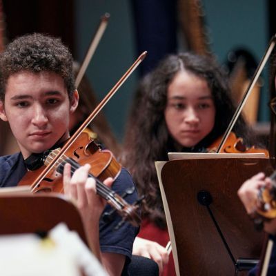 Three Junior Guildhall students on stage playing the violin concentrating and reading sheet music